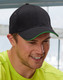 A black cap with a green peak, featuring a logo, worn by a smiling man in a bright yellow shirt.