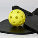 A yellow pickleball with holes, resting on a black paddle and a round black surface, featuring a logo.