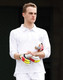 A white men's 3/4 sleeve cricket polo with a logo, worn by a model holding a cricket bat and gloves.