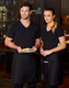 Two individuals wear black short waist aprons over black tops, standing with a plate of food in a restaurant setting.