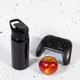 A drink bottle in glossy black with a free flow lid, alongside a red apple and a gaming controller.