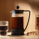 A Bodum Brazil French Press in black with a glass body, sitting next to a glass cup of coffee on a wooden shelf.