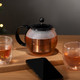 A glass tea press with a black handle, filled with brewed tea, alongside two cups and a smartphone on a wooden surface.