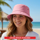 A pink bucket hat sits on a smiling woman's head with a beach backdrop and palm trees. The hat features a logo.