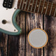 A round white Bluetooth speaker with a wooden base, placed beside a teal electric guitar on a rustic wooden surface.