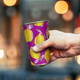 A drink cup featuring a vibrant purple design with yellow lemons and a silver rim, held in a hand.