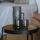 A drink bottle and two tumblers in smoky grey, placed on a round table beside a couch. The items feature a sleek design.