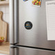 A round black magnetic badge with a logo attached to a stainless steel fridge, alongside a notepad and groceries.