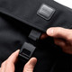 A black messenger bag with a flap and fastening mechanism, featuring a logo. A hand is holding the clasp.