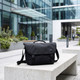 A black satchel bag sits on a concrete bench in a modern urban setting with greenery. It features a logo.