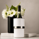 A white wine bottle cooler with a silver band, placed on a marble surface beside a black vase with white flowers.