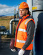 A man wearing an orange hi-vis hooded puffer vest stands beside a metallic structure, holding a laptop.
