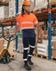 A man wearing orange and navy work pants with reflective tapes, accompanied by a hard hat and boots, stands in a warehouse.