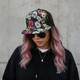A floral-patterned flat peak mesh cap in black with bright pink and yellow flowers, worn by a person with pink hair.