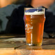 A transparent beer glass filled with amber-coloured beer, featuring a logo and set on a wooden table.