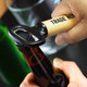 A drink bottle opener with a wooden handle and a black metal opener, being used to open a bottle.