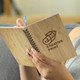 A medium bamboo notebook with a spiral binding, held by someone writing with a pen. It features a printed logo.