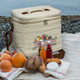A beige cooler bag with a logo, surrounded by fruits, glass bottles, and bread on a textured surface.