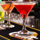 A vibrant red cocktail in a martini glass on a black PVC bar mat, with another drink in the background.