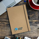 A brown hardcover notebook with an elastic band, featuring a logo, placed on a wooden desk alongside a coffee cup and stationery. A brown hardcover notebook with an elastic band, featuring a logo, placed on a wooden desk alongside a coffee cup and stationery.