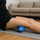 A blue branded massage ball positioned on a yoga mat with a person’s leg resting on it, in a living room setting.