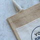 A jute tote bag featuring a natural beige colour with a cream panel and a decorative logo. A jute tote bag featuring a natural beige colour with a cream panel and a decorative logo.