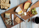 A wooden cheese board featuring various cheeses, crackers, grapes, almonds, and a set of six silver cheese knives.
