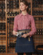 A denim half apron with a beige tie, worn by a model in a checked shirt, set against a wine rack background.
