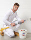 A model kneels in white utility stretch cargo work pants, holding a paint roller and bucket, wearing tan work boots.