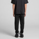 Black youth track pants are worn with a matching dark shirt and black shoes, showcased from the back.