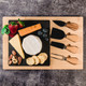 A bamboo cheese board featuring assorted cheeses, fruits, nuts, and four wooden handled cheese knives.