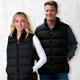 Two individuals wearing black puffa vests, standing against a textured white background. The vests have front zip closures and pockets.