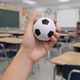 A hand holds a white and black soccer ball stress reliever in a classroom setting with desks and a chalkboard.