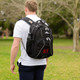 A black backpack with multiple compartments and a logo, worn by a person outdoors in a grassy area.