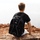 A black High Sierra Swerve 17" backpack with white accents is worn by a person sitting on a rocky surface by the ocean.