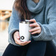 A drink bottle featuring a white exterior with a black logo, being held by a person in a grey sweater.