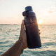 A drink bottle in dark blue with a lid, held against a sunset over a body of water, featuring a logo.