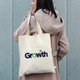 A cream tote bag with sturdy handles, featuring a logo on one side, carried by a person in a light coat.
