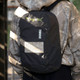 A black Thule Achiever 15 laptop backpack with a logo, worn by a person in a camouflage hoodie.