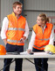 Two individuals in high-visibility orange safety vests with 3M reflective tapes, standing by a table with tools and helmets.