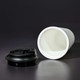 A white coffee mug made from recycled materials, featuring a black lid, displayed on a dark background.