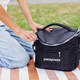 A black cooler bag with a zippered top and mesh pocket, sitting on a striped blanket. It has a logo.