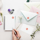 A coloured round stamp in purple, placed alongside envelopes, a notepad, flowers, and a pen on a table.