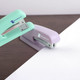 A mint green and light purple stapler on a wooden surface, with a sheet of white paper beside it.