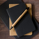 A cardboard pen rests atop two notebooks, one black and one tan, on a wooden surface.