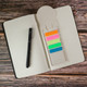 A circle-shaped bookmark in neutral tones with vibrant coloured sections resting on an open notebook beside a pen.