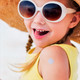 A child in sunglasses and a sun hat smiles, with a small sticker on their arm, wearing a yellow top.