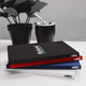 A set of three Pioneer RPET A5 Notebooks in black, red, and white, placed beside a plant and a drink.