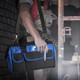 A tool bag in black and blue with multiple pockets, held by a person wearing work attire. It has a logo on the front.