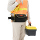 A waist tool bag in black with red accents, worn by a person holding a yellow tool box and wearing a high-visibility vest.